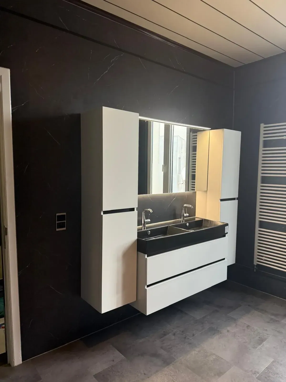 Badkamer met donkere marmerlook wandpanelen, zwevend wit meubel met dubbele zwarte wastafel, ledspiegel en kolomkasten