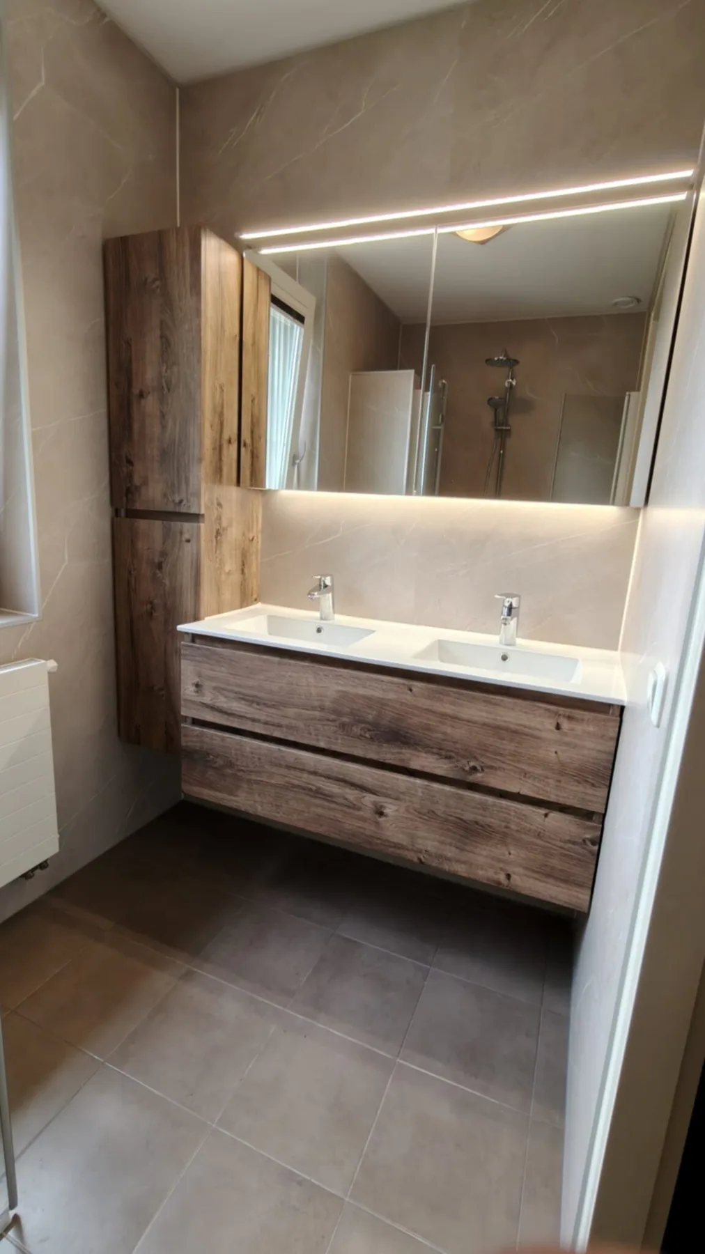 Dubbel wastafelmeubel in houtlook met ledspiegel en hoge kast, beige marmerlook wandpanelen en grijze vloer