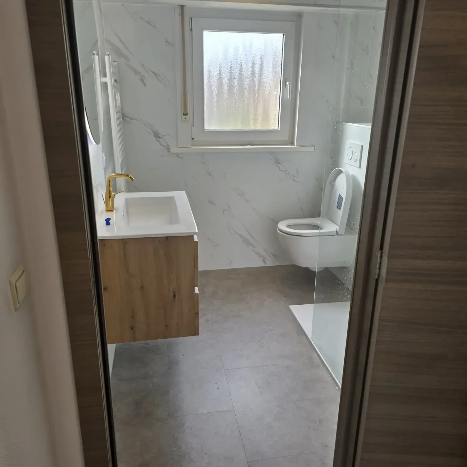 Doorkijk naar een badkamer met marmerlook wandpanelen, eiken wastafelmeubel met gouden kraan en hangtoilet