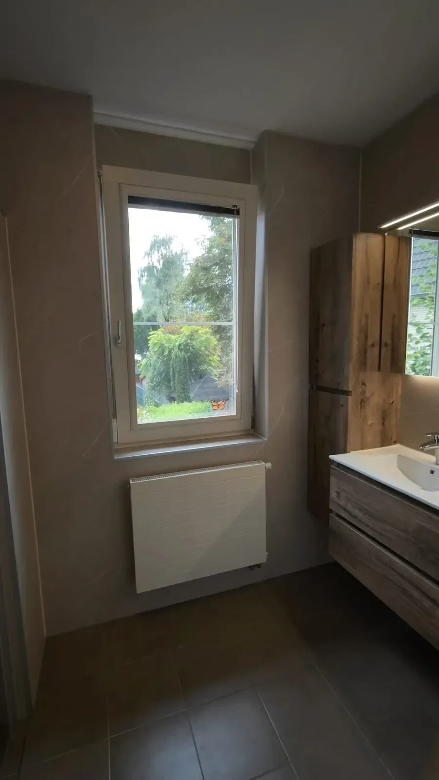 Badkamer met raam en radiator, wastafelmeubel en hoge kast in houtlook, donkere vloertegels