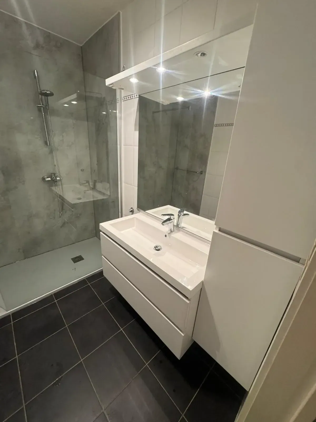 Badkamer met wit wastafelmeubel, spiegelkast met verlichting en inloopdouche met grijze wandpanelen