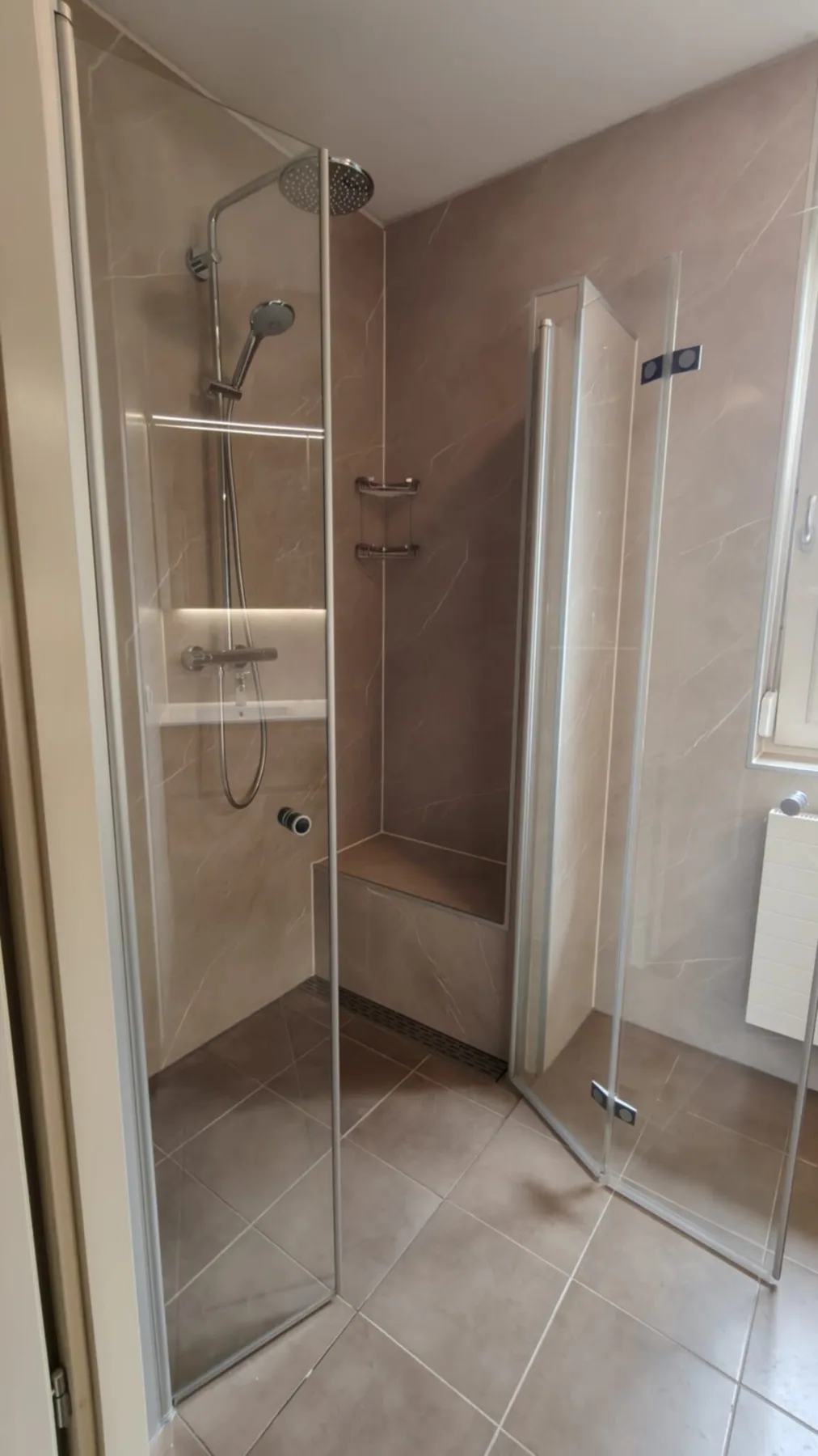 Douche met glazen vouwdeuren, regendouche, nis en zitbankje tegen beige marmerlook panelen