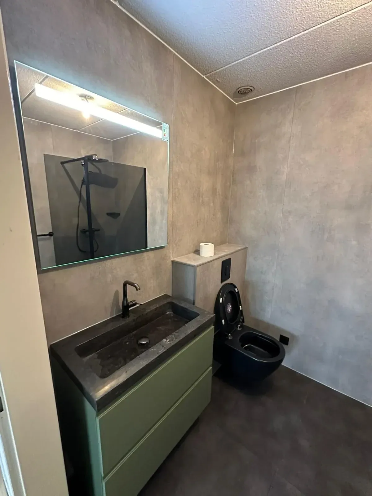 Groen wastafelmeubel met natuurstenen wasbak, ledspiegel en zwart hangtoilet tussen grijze wandpanelen