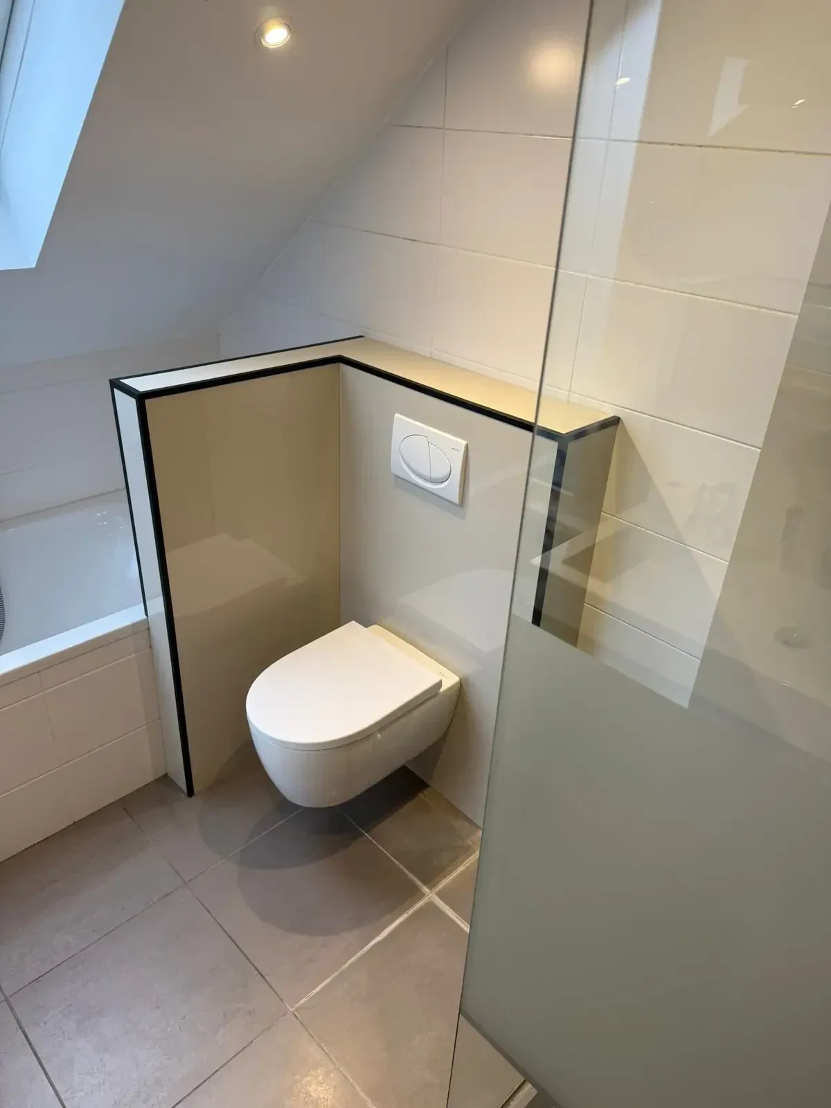 Hangtoilet op voorzetwand met zwarte afwerkrand onder schuin dak, naast ligbad en glazen wand