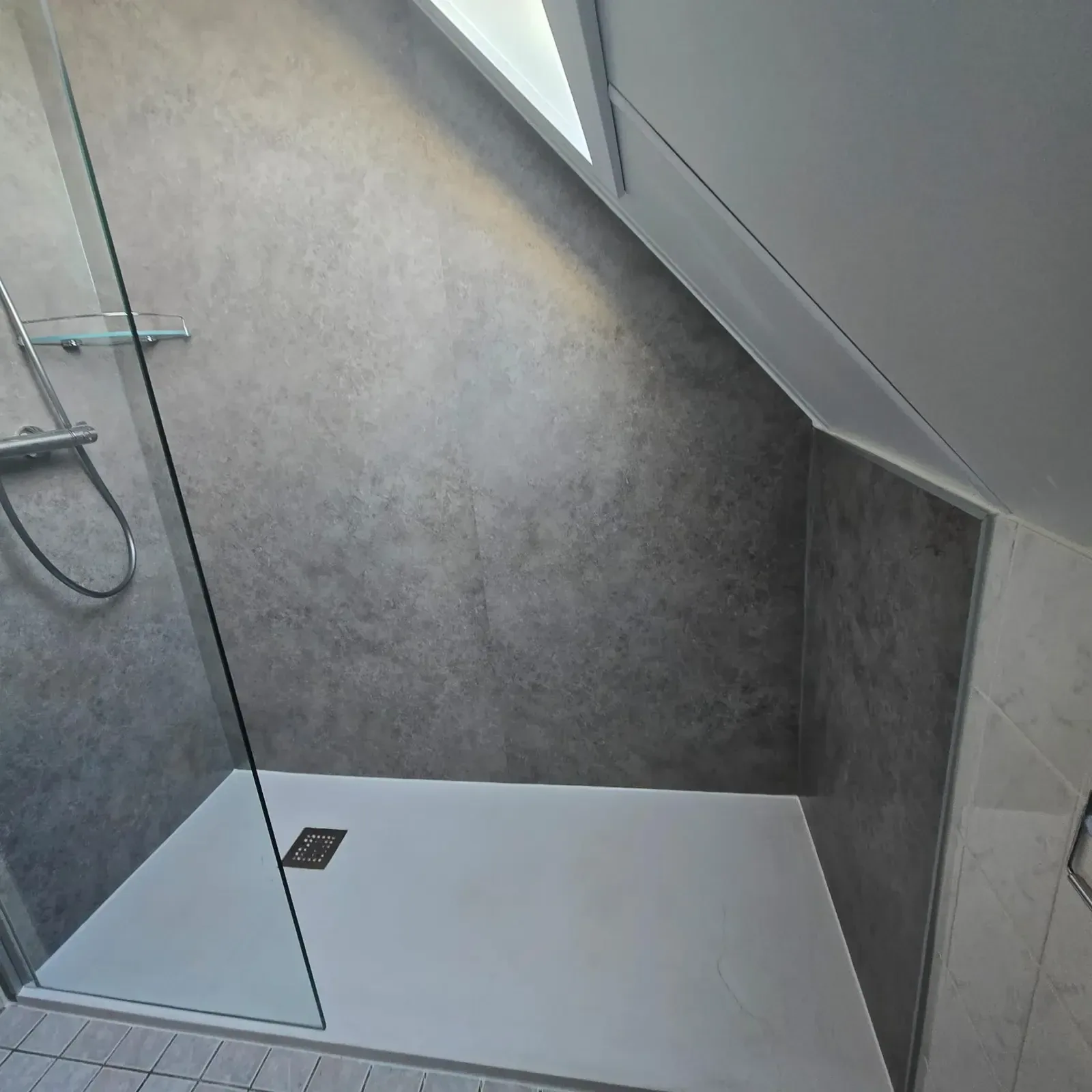 Inloopdouche onder schuin dak met grijze betonlook wandpanelen, glazen wand en witte douchebak