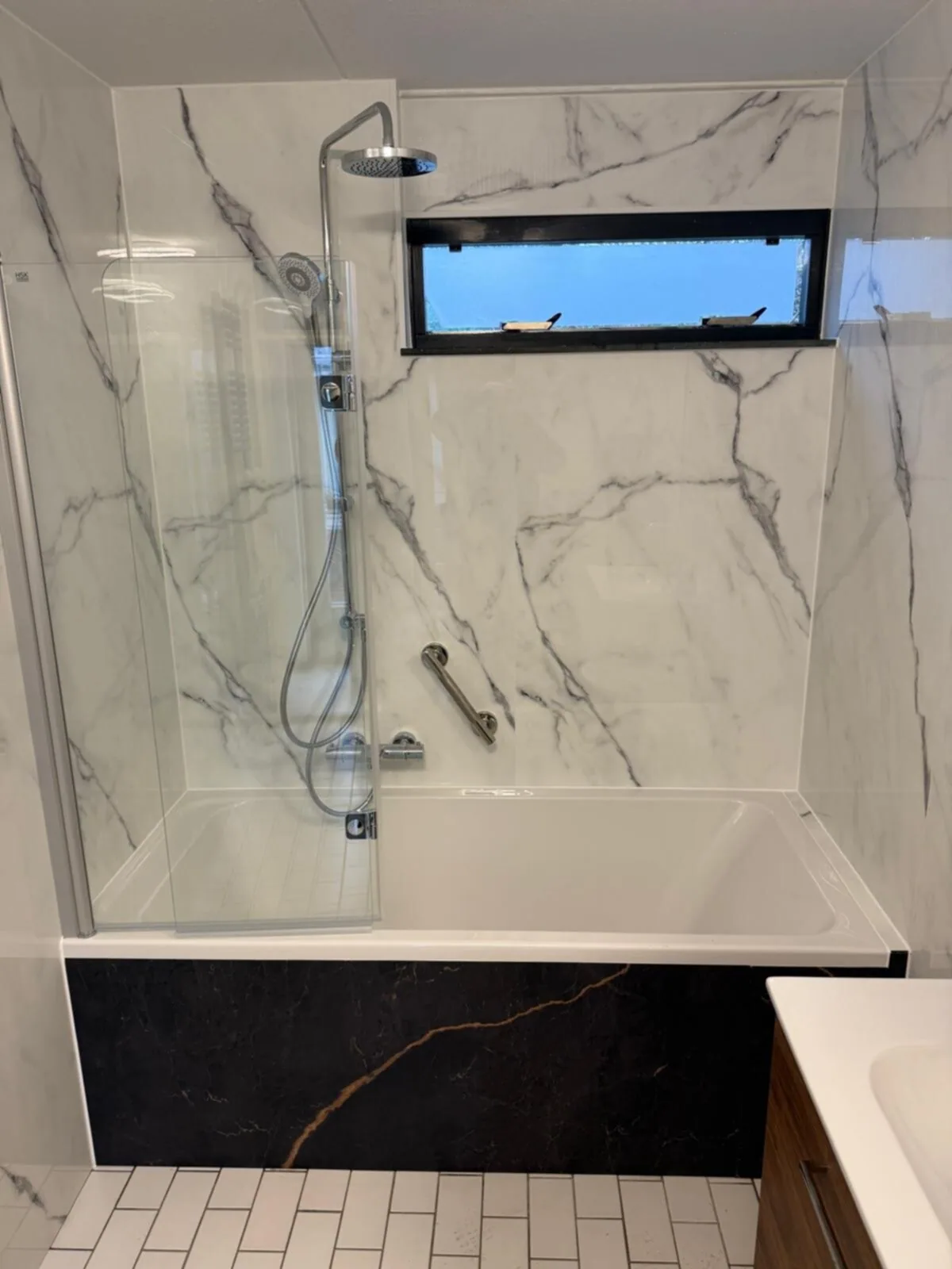 Ligbad met badwand en regendouche tegen witte marmerlook wandpanelen, zwarte marmerlook ombouw