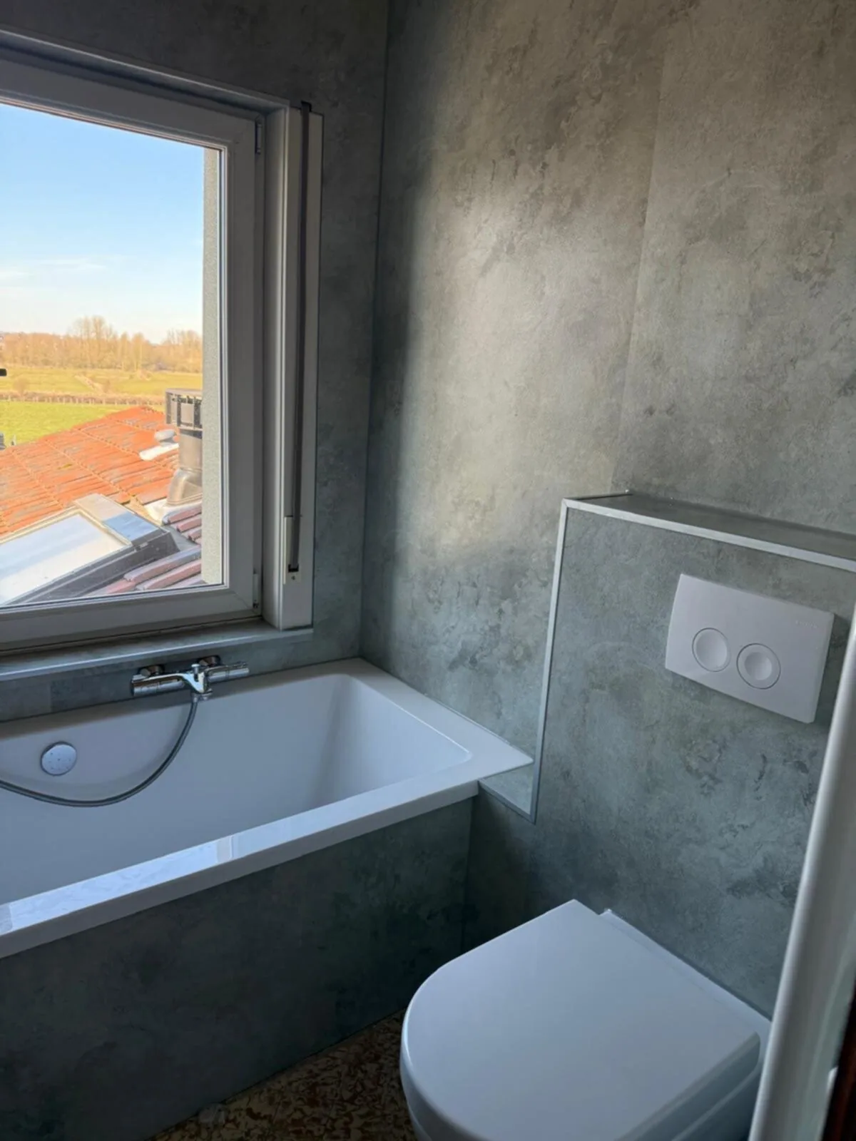 Ligbad onder het raam met uitzicht, grijze betonlook wandpanelen en hangtoilet