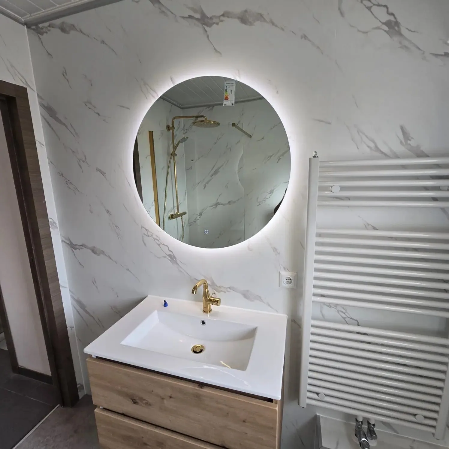 Ronde ledspiegel boven eiken wastafelmeubel met gouden kraan tegen marmerlook wandpanelen