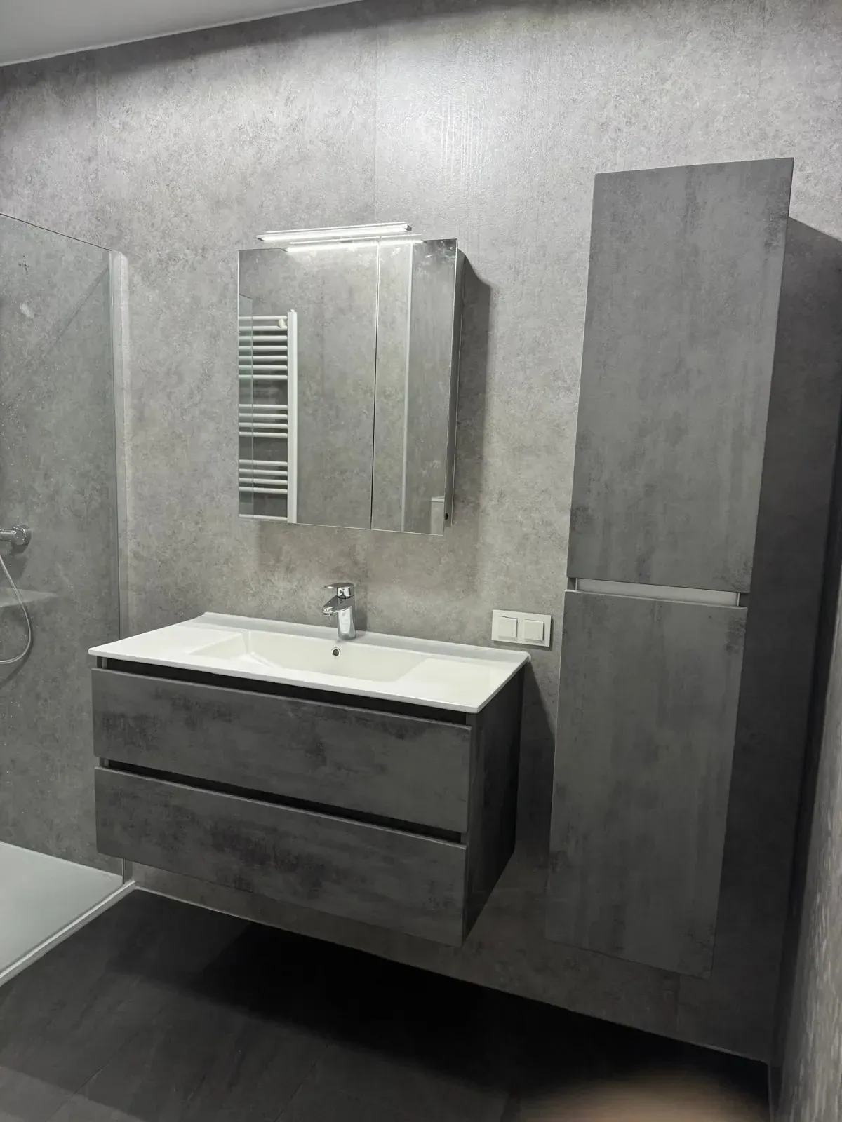 Grijs betonlook wastafelmeubel met spiegelkast en hoge kolomkast tegen grijze steenlook wandpanelen