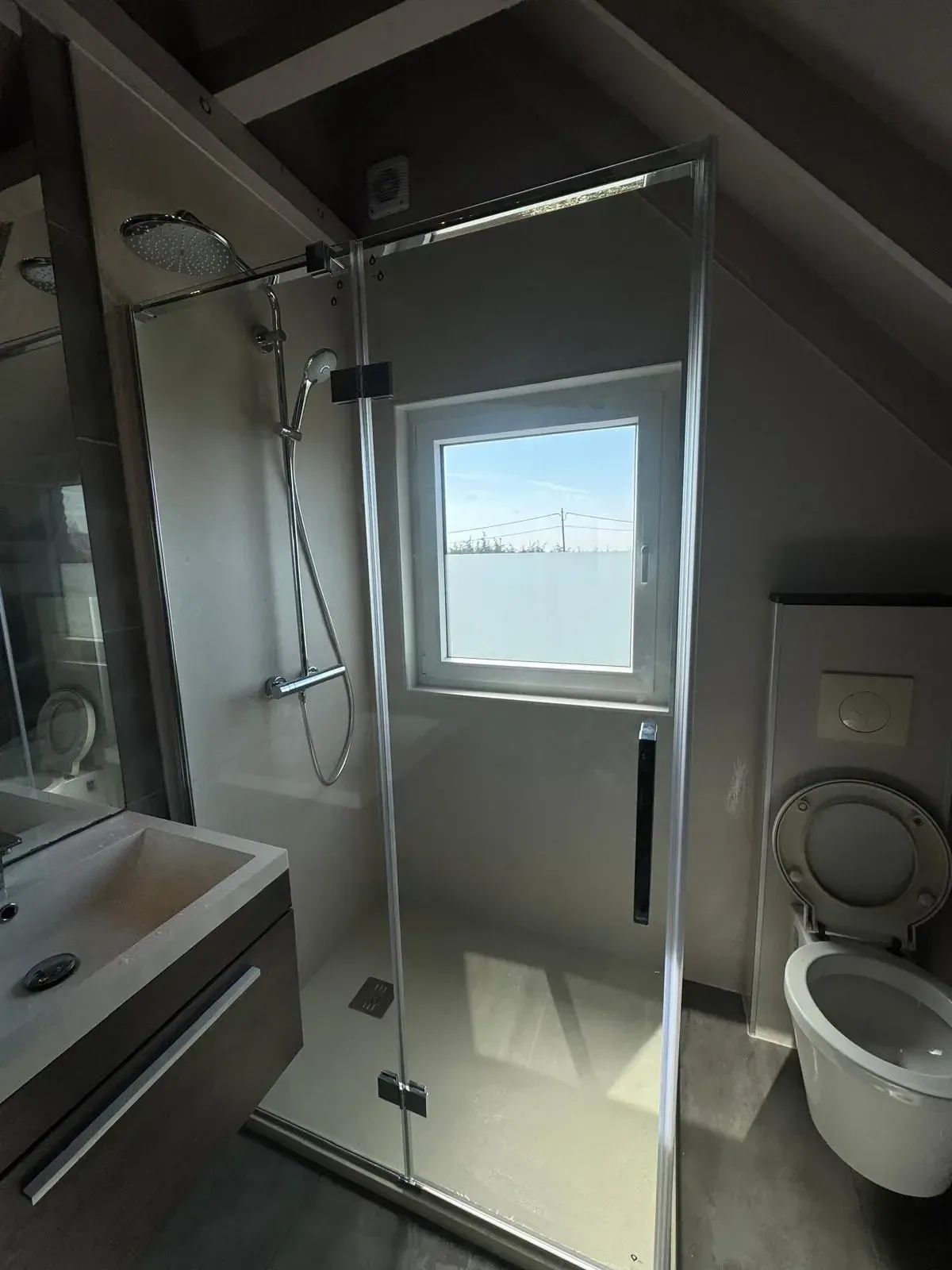 Zolderbadkamer met glazen douchecabine onder schuin dak, hangtoilet en wastafelmeubel